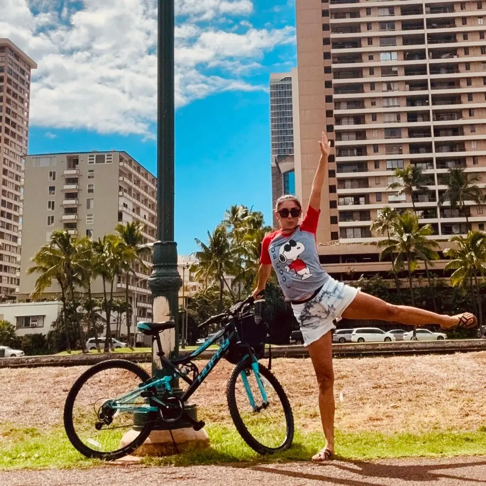 Daniela kicking a leg up beside her bike in Honolulu, palm trees and towers behind her